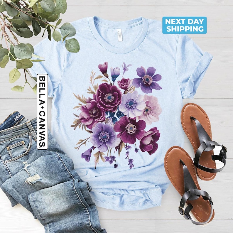 May include: Light blue t-shirt with a floral design in shades of purple and white. The shirt has the text "BELLA + CANVAS" on the side. A pair of brown sandals with black straps are next to the shirt.