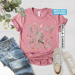 May include: A pink t-shirt with a floral print featuring butterflies and leaves. The shirt has a crew neck and short sleeves. The text "NEXT DAY SHIPPING" and "BELLA + CANVAS" are visible on the image.