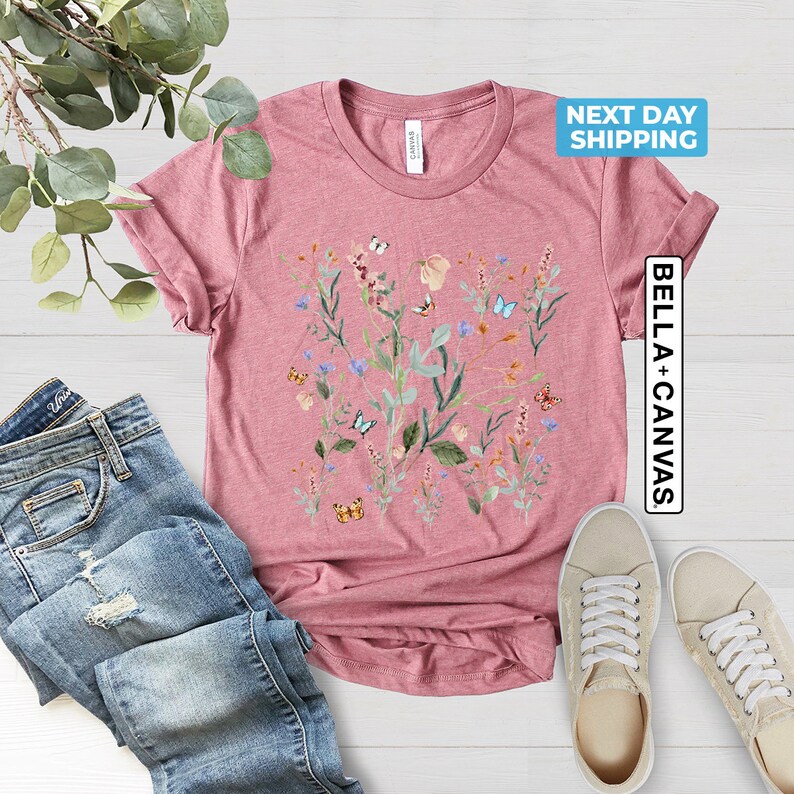 May include: A pink t-shirt with a floral print featuring butterflies and leaves. The shirt has a crew neck and short sleeves. The text "NEXT DAY SHIPPING" and "BELLA + CANVAS" are visible on the image.