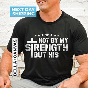 May include: Black t-shirt with white text that reads "Not by my strength but his" with a cross and stars above the text.