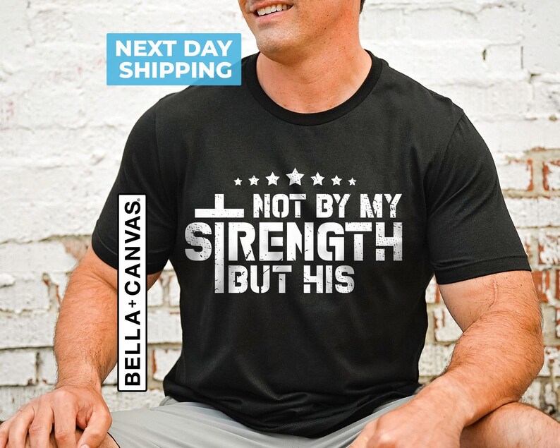 May include: Black t-shirt with white text that reads "Not by my strength but his" with a cross and stars above the text.