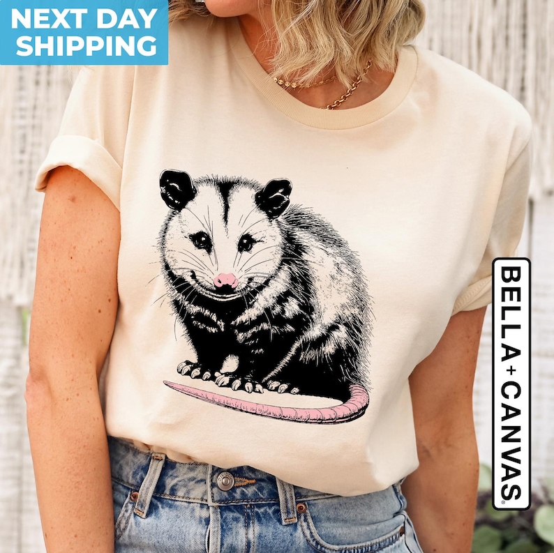 K&ouml;nnte beinhalten: Ein cremefarbenes T-Shirt mit einer schwarz-wei&szlig;en Illustration eines Opossums. Das Opossum l&auml;chelt und hat eine rosa Nase. Der Text "BELLA + CANVAS" ist auf der rechten Seite des Shirts aufgedruckt.
