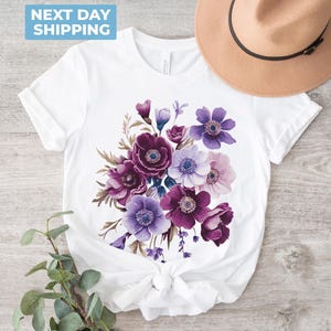 May include: White t-shirt featuring a watercolor floral design in shades of purple, lavender, and white. The bouquet includes anemones and other flowers. A tan hat with a black band rests on the shirt. The shirt is tied at the waist.