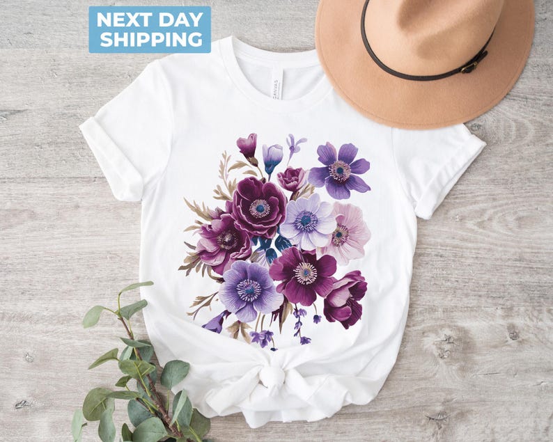 May include: White t-shirt featuring a watercolor floral design in shades of purple, lavender, and white. The bouquet includes anemones and other flowers. A tan hat with a black band rests on the shirt. The shirt is tied at the waist.