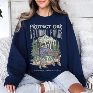 May include: Navy blue sweatshirt with a graphic that reads "PROTECT OUR NATIONAL PARKS." The graphic features a mountain range, trees, a river, and a fish. The text "EXPLORE RESPONSIBLY" is below the graphic.