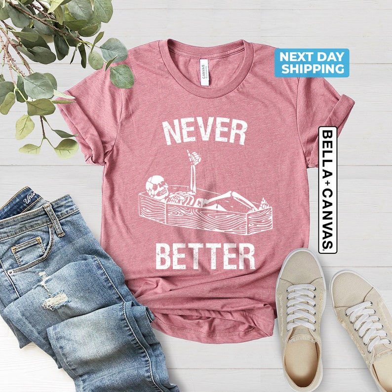 May include: A pink t-shirt with a white graphic of a skeleton lying in a coffin with the text "NEVER BETTER".