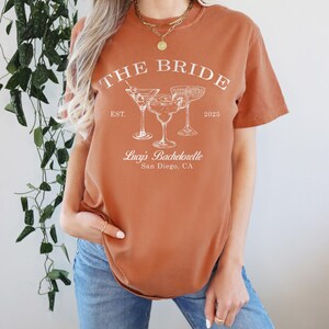 May include: A rust-colored t-shirt with the text "THE BRIDE" in white, with the dates "EST. 2025" and the text "Lucy's Bachelorette San Diego, CA" below. The shirt features a graphic of three cocktail glasses.