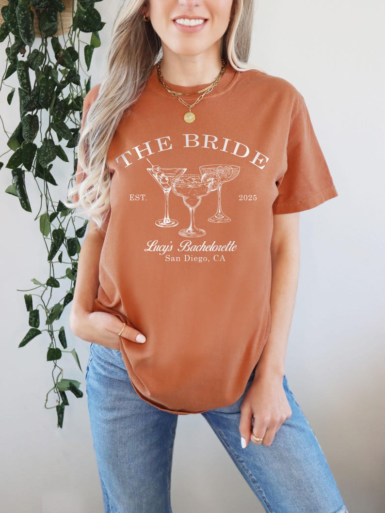 May include: A rust-colored t-shirt with the text "THE BRIDE" in white, with the dates "EST. 2025" and the text "Lucy's Bachelorette San Diego, CA" below. The shirt features a graphic of three cocktail glasses.