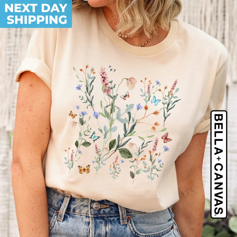 May include: A beige t-shirt with a floral print featuring blue, pink, and green flowers and butterflies. The text "BELLA + CANVAS" is printed on the right side of the shirt.