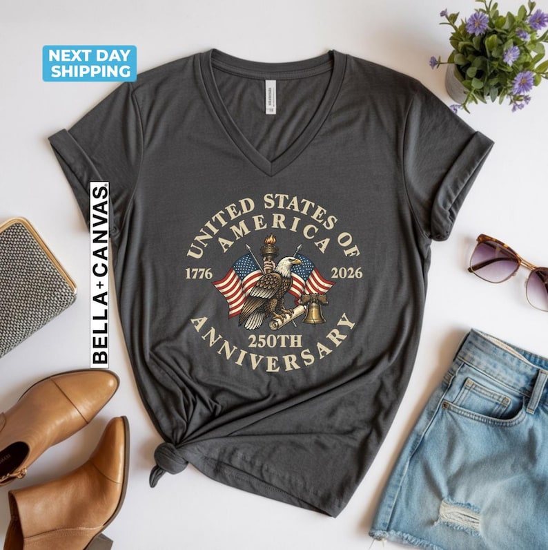 May include: Dark gray V-neck t-shirt with a graphic commemorating the 250th anniversary of the United States of America. The design includes an eagle, flags, and the text "United States of America 250th Anniversary."