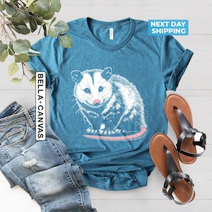 K&ouml;nnte beinhalten: Ein blaues T-Shirt mit einem wei&szlig;en Aufdruck eines Opossums. Das Opossum blickt auf den Betrachter und hat eine rosafarbene Nase. Der Text "BELLA + CANVAS" befindet sich auf der linken Seite des Shirts.