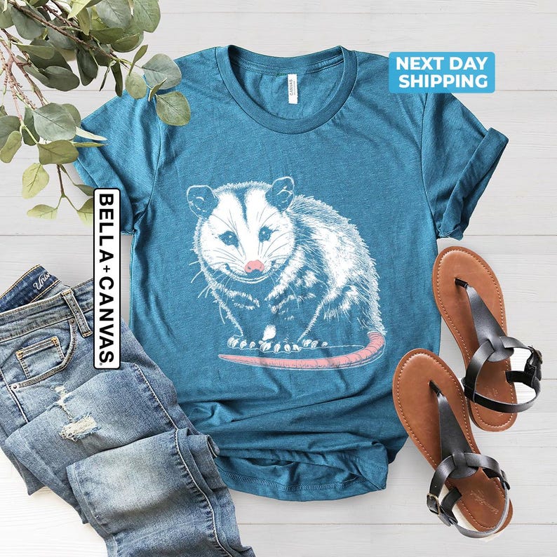 K&ouml;nnte beinhalten: Ein blaues T-Shirt mit einem wei&szlig;en Aufdruck eines Opossums. Das Opossum blickt auf den Betrachter und hat eine rosafarbene Nase. Der Text "BELLA + CANVAS" befindet sich auf der linken Seite des Shirts.