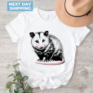 K&ouml;nnte beinhalten: Wei&szlig;es T-Shirt mit einer schwarz-wei&szlig;en Illustration eines Opossums mit rosa Nase. Das Opossum sitzt auf seinen Hinterbeinen, die Schwanz um seinen K&ouml;rper gewickelt.