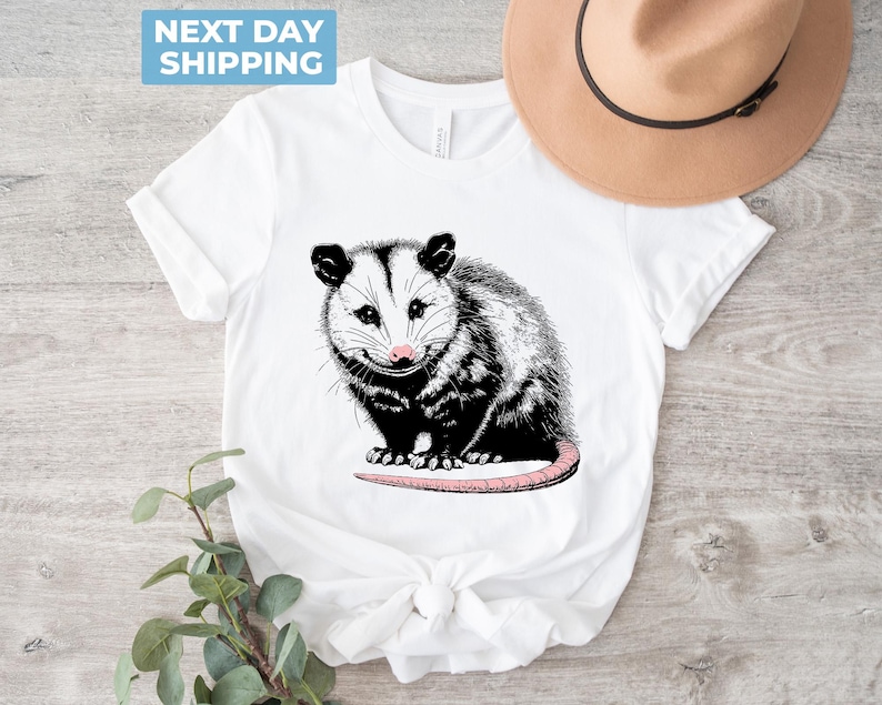 K&ouml;nnte beinhalten: Wei&szlig;es T-Shirt mit einer schwarz-wei&szlig;en Illustration eines Opossums mit rosa Nase. Das Opossum sitzt auf seinen Hinterbeinen, die Schwanz um seinen K&ouml;rper gewickelt.