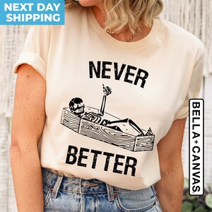 May include: A beige t-shirt with a black graphic of a skeleton in a coffin with the text "NEVER BETTER" printed on the front.