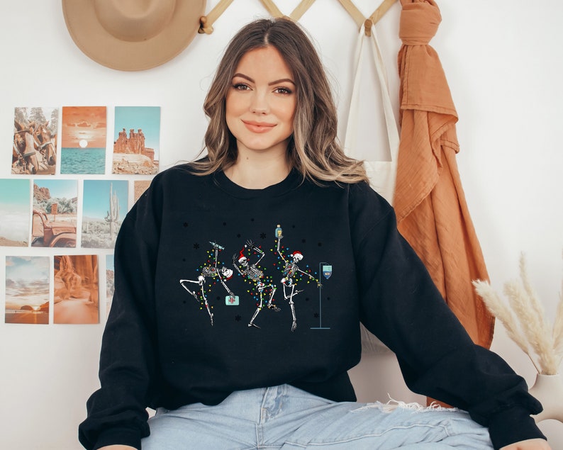 May include: A black sweatshirt with a festive design featuring three skeletons wearing Santa hats and holding drinks. The skeletons are decorated with colorful lights.
