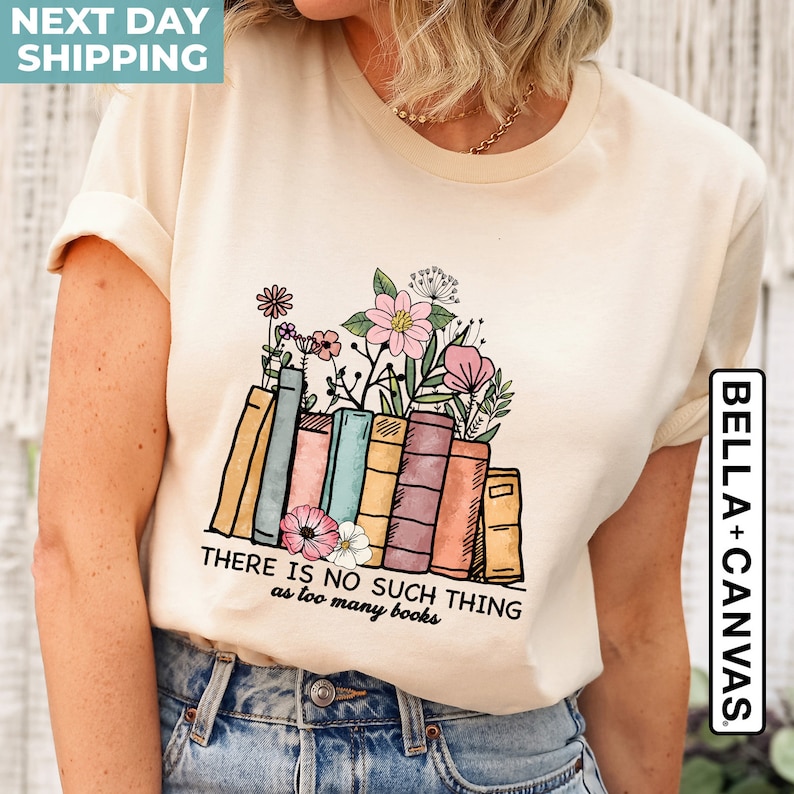 May include: A beige t-shirt with a graphic of a stack of colorful books with flowers growing out of the top. The text "THERE IS NO SUCH THING as too many books" is printed below the graphic. The shirt is a Bella + Canvas brand.