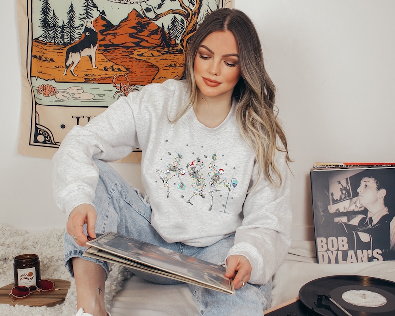 May include: A gray sweatshirt with a festive design of skeletons dressed as Christmas trees. The skeletons are holding drinks and wearing Santa hats. The sweatshirt is being worn by a person sitting on a rug with a record player and vinyl records.