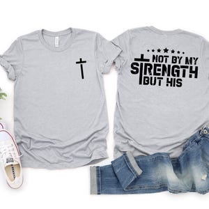 May include: A gray t-shirt with a cross on the front and the text "Not by my strength but his" on the back.