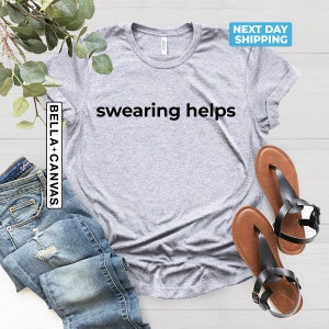 Op de afbeelding: Een grijs T-shirt met de tekst "swearing helps" in zwart gedrukt op de voorkant. Het shirt ligt plat op een wit oppervlak met een paar bruine sandalen en een blauwe spijkerbroek.