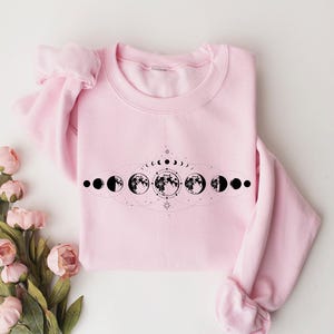 May include: A pink sweatshirt with a black graphic of a moon phase design. The design features a crescent moon, a half moon, a full moon, and a waning gibbous moon.
