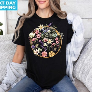 May include: Black t-shirt with a floral design in a circle. The flowers are pink, yellow, blue, and white. The circle is outlined in gold.
