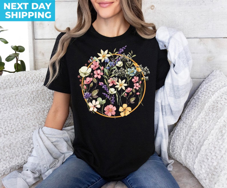 May include: Black t-shirt with a floral design in a circle. The flowers are pink, yellow, blue, and white. The circle is outlined in gold.