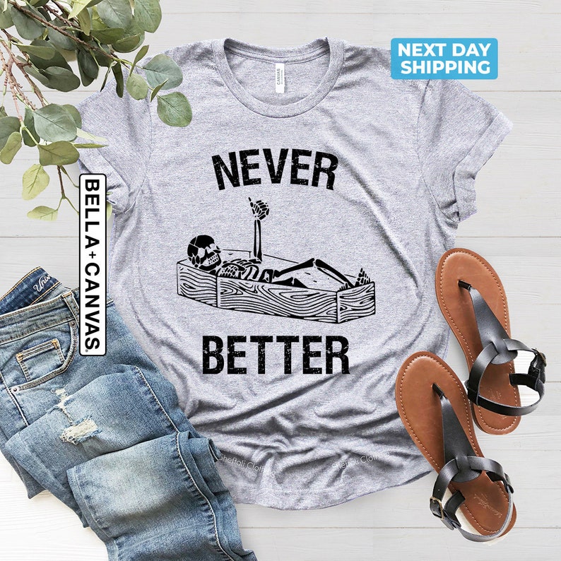 May include: A gray t-shirt with the text "NEVER BETTER" printed on it. A skeleton is lying in a coffin with its arm raised in a thumbs up gesture. The text "BELLA + CANVAS" is printed on the left side of the shirt.