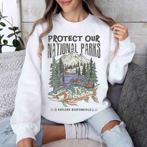 May include: White sweatshirt with a graphic that reads "PROTECT OUR NATIONAL PARKS." The graphic features a mountain range, trees, a river, and a fish. The text "EXPLORE RESPONSIBLY" is below the graphic.