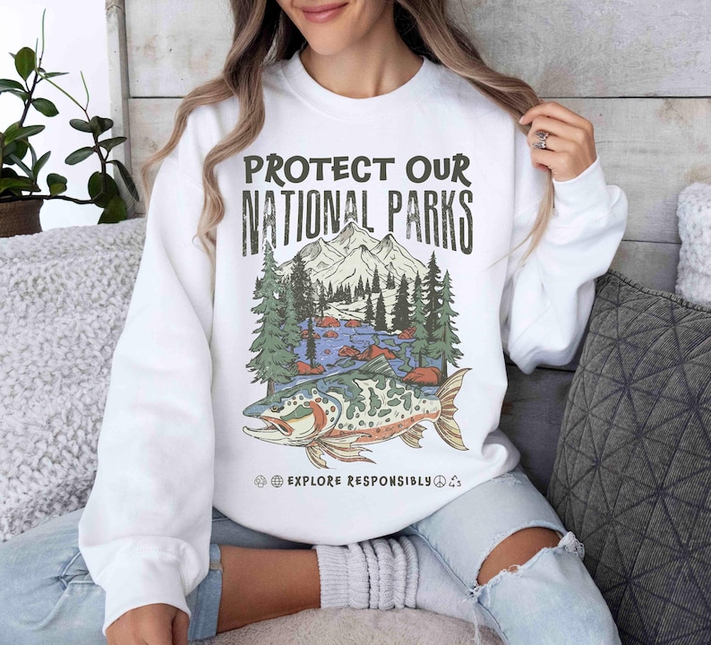 May include: White sweatshirt with a graphic that reads "PROTECT OUR NATIONAL PARKS." The graphic features a mountain range, trees, a river, and a fish. The text "EXPLORE RESPONSIBLY" is below the graphic.