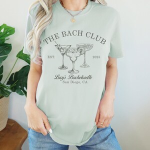 May include: A light green t-shirt with the text "The Bach Club Est. 2025" and a graphic of three cocktail glasses. The text "Lucy's Bachelorette San Diego, CA" is also printed on the shirt.