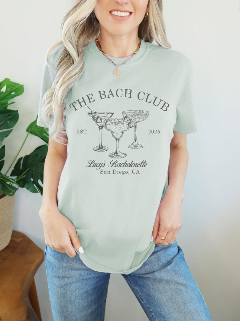 May include: A light green t-shirt with the text "The Bach Club Est. 2025" and a graphic of three cocktail glasses. The text "Lucy's Bachelorette San Diego, CA" is also printed on the shirt.