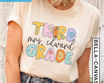 May include: A light beige t-shirt with the text "THIRD GRADE" in colorful letters. The text is decorated with small illustrations of school supplies, including a pencil, a ruler, a globe, and a star. The text "mrs. edward" is written in black below the main text.