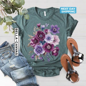 May include: Teal t-shirt with a floral design featuring purple, violet, and white anemone flowers. The shirt is paired with blue jeans and brown sandals. The text "BELLA + CANVAS" is printed vertically on the left side of the shirt.