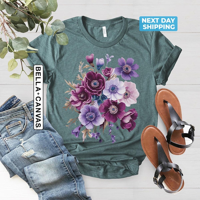 May include: Teal t-shirt with a floral design featuring purple, violet, and white anemone flowers. The shirt is paired with blue jeans and brown sandals. The text "BELLA + CANVAS" is printed vertically on the left side of the shirt.