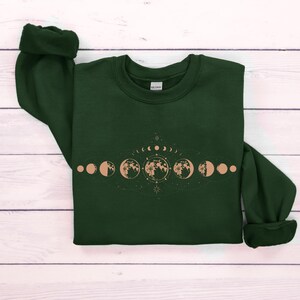 May include: A dark green sweatshirt with a graphic of a moon phase cycle in a light brown color. The graphic includes a crescent moon, half moon, full moon, and other phases of the moon.