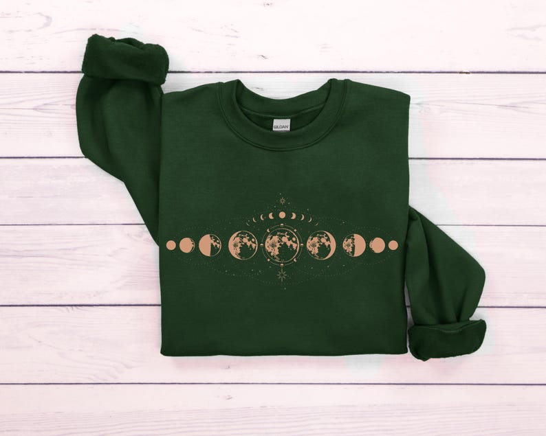 May include: A dark green sweatshirt with a graphic of a moon phase cycle in a light brown color. The graphic includes a crescent moon, half moon, full moon, and other phases of the moon.