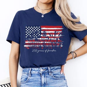 May include: Navy blue t-shirt featuring a distressed American flag design with red and white stripes, white stars, and wheat stalks. The text "250 years of freedom" is printed below the flag. The shirt is a classic crew neck.