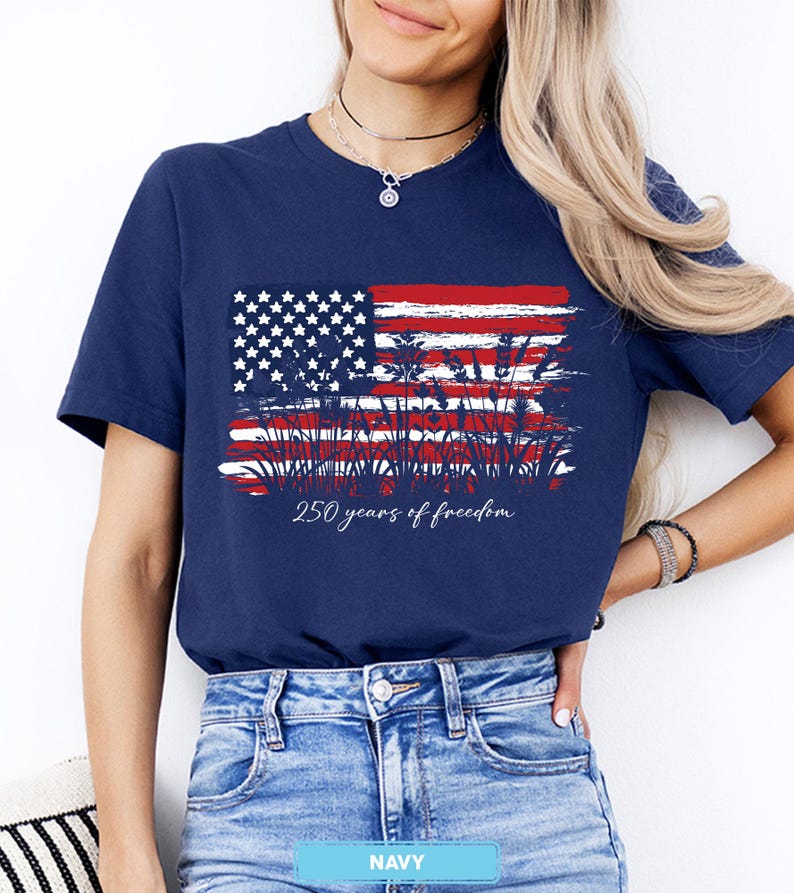 May include: Navy blue t-shirt featuring a distressed American flag design with red and white stripes, white stars, and wheat stalks. The text "250 years of freedom" is printed below the flag. The shirt is a classic crew neck.