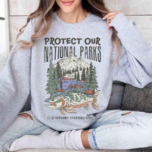 May include: A light gray sweatshirt with the text "PROTECT OUR NATIONAL PARKS" above a graphic of a mountain scene with trees, water, and a fish. The words "EXPLORE RESPONSIBLY" are below the graphic.