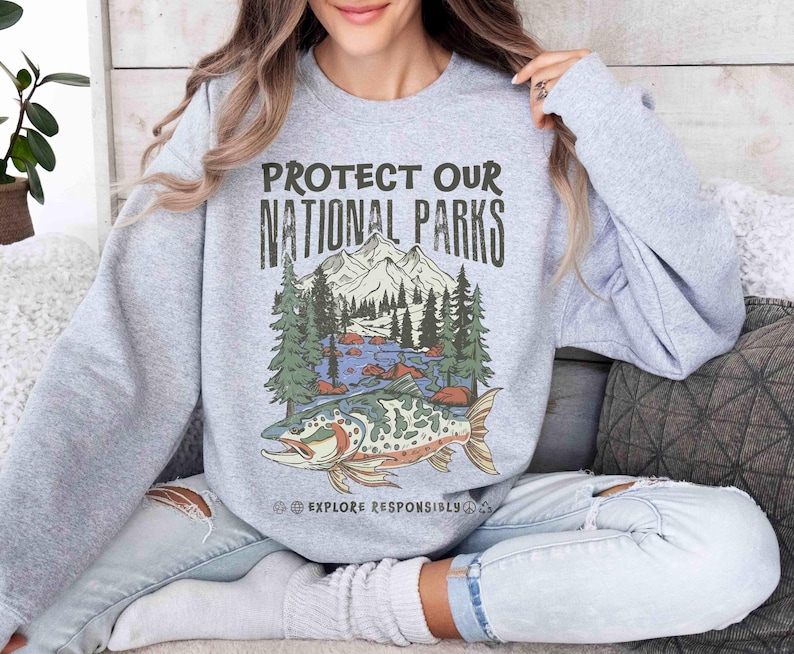 May include: A light gray sweatshirt with the text "PROTECT OUR NATIONAL PARKS" above a graphic of a mountain scene with trees, water, and a fish. The words "EXPLORE RESPONSIBLY" are below the graphic.