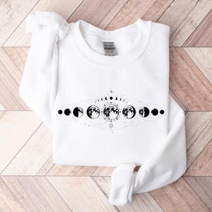 May include: White sweatshirt with a black and white moon phases design. The design features a crescent moon, half moon, full moon, and other phases of the moon.