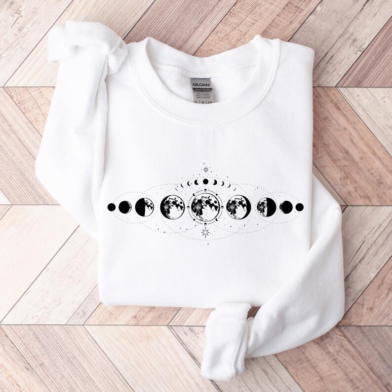 May include: White sweatshirt with a black and white moon phases design. The design features a crescent moon, half moon, full moon, and other phases of the moon.