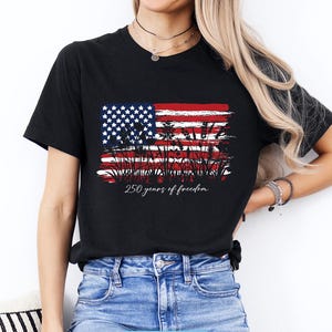 May include: Black t-shirt featuring a distressed American flag design with red and white stripes, blue stars, and wheat stalks. The text "250 years of freedom" is printed below the flag. The shirt is made of a soft material.