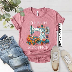 May include: Dusty rose t-shirt featuring a sewing machine design with the text "I'll Be In My Office." The design includes spools of thread, a quilt, and a floral pattern. The shirt is paired with blue jeans and sneakers.