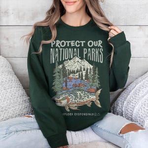May include: Dark green sweatshirt with the text "PROTECT OUR NATIONAL PARKS" above a graphic of a mountain scene, trees, and a fish. The words "EXPLORE RESPONSIBLY" are below the graphic. The sweatshirt is a long-sleeved crew neck.