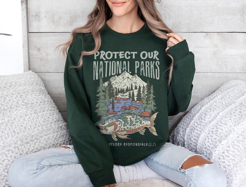 May include: Dark green sweatshirt with the text "PROTECT OUR NATIONAL PARKS" above a graphic of a mountain scene, trees, and a fish. The words "EXPLORE RESPONSIBLY" are below the graphic. The sweatshirt is a long-sleeved crew neck.