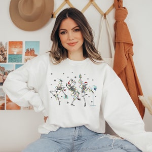 May include: White sweatshirt with a Christmas-themed design featuring three skeletons dressed as Christmas trees with lights and ornaments. The skeletons are holding drinks and dancing.