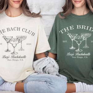 May include: Two white t-shirts with a bachelorette party theme. The text on the shirts reads "The Bach Club" and "The Bride". Both shirts feature a graphic of three cocktail glasses with olives and a text that reads "Lucy's Bachelorette, San Diego, CA".
