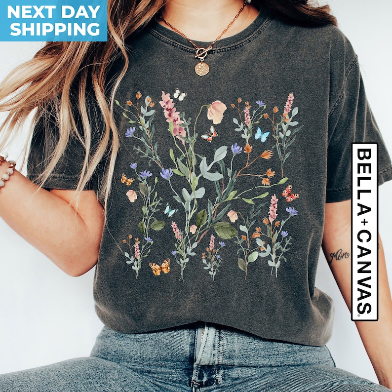 May include: A dark grey t-shirt with a floral print featuring pink, blue, and green flowers and butterflies. The text "Bella + Canvas" is printed on the right side of the shirt.